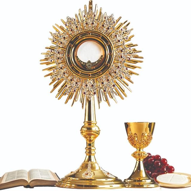 A gold monstrance with the consecrated Host, a chalice, grapes, and an open Bible — the symbols of the Eucharist